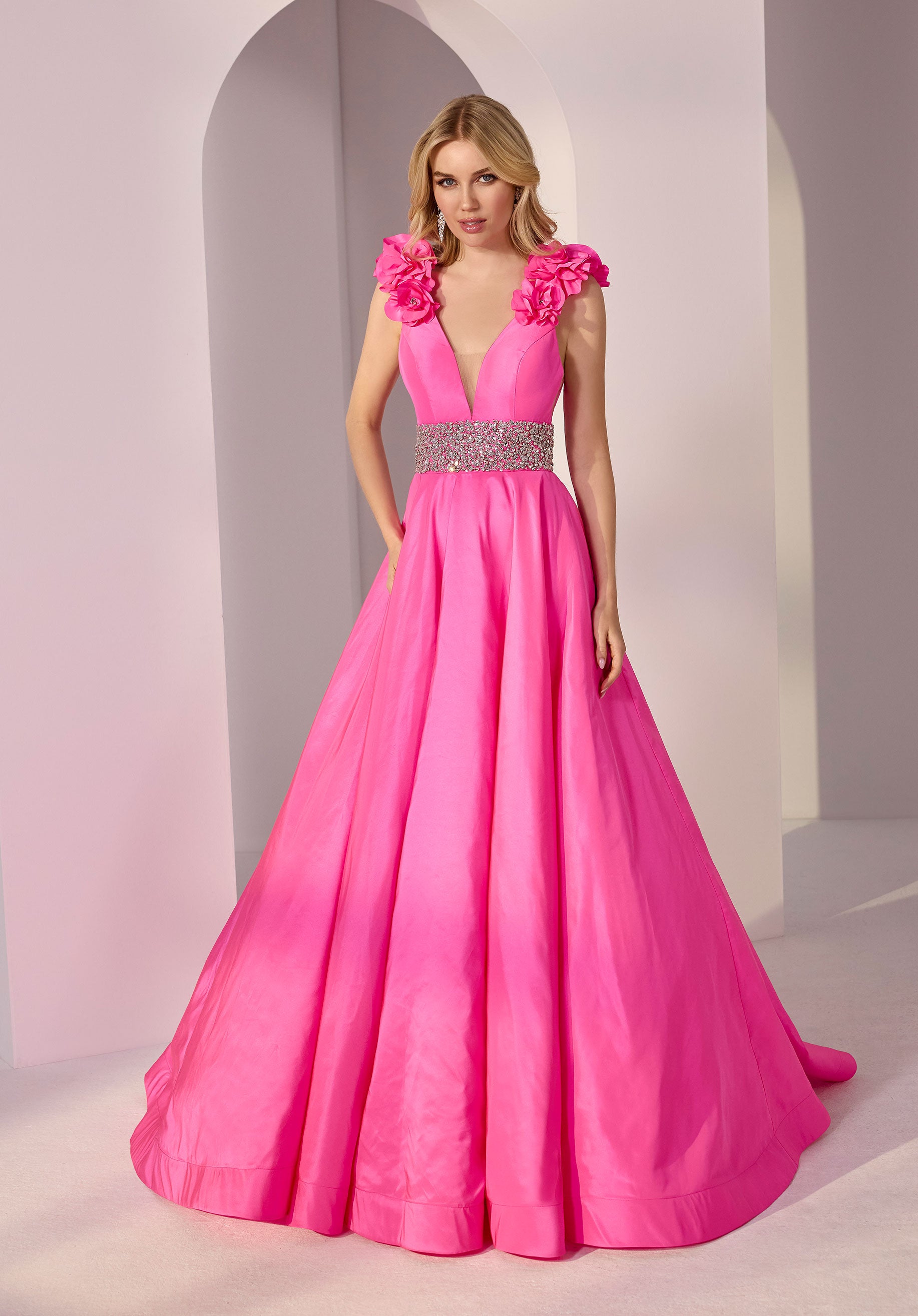 A woman stands in a light, arched setting wearing the Long Prom Dress: Morilee 49202 in fuchsia, featuring 3D rosettes on the shoulders, a deep V neckline, and a jeweled waistband.